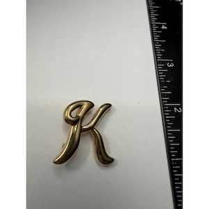 Puede incluir: Broche de metal dorado con forma de la letra "K". El broche tiene un estilo de fuente cursiva y mide aproximadamente 3,8 cm de altura. El broche está sobre una superficie blanca.
