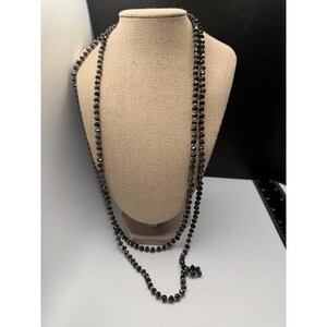 May include: A long, layered necklace with small, dark beads. The necklace is draped over a tan mannequin bust, showcasing its length and design. The beads appear to be a dark, possibly black or charcoal colour, with a subtle sheen.