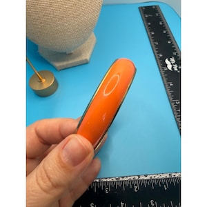 Puede incluir: Brazalete de color naranja brillante con un borde de metal dorado. El brazalete se sostiene en una mano sobre un fondo azul. También se ven una regla y un expositor de joyas.