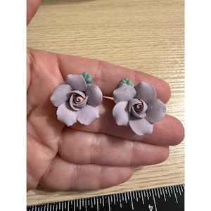 May include: Two handmade lavender flower earrings. Each earring features a detailed flower with multiple petals, a dark pink center, and small green leaves. The earrings are displayed on a light-colored surface.