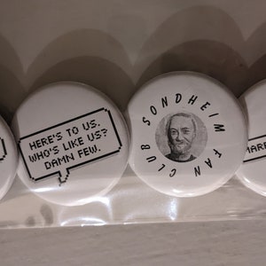 May include: Four white round buttons with black text and graphics. One button says "ART ISN'T EASY" in a speech bubble. Another says "HERE'S TO US. WHO'S LIKE US? DAMN FEW." A third button has a portrait and says "SONDHEIM FAN CLUB." The last says "MARRY ME A LITTLE."