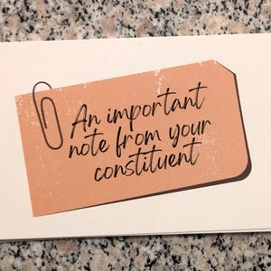 May include: A stack of white notecards with a peach-colored tag featuring the text "An important note from your constituent" in black script. A black paperclip is attached to the tag.