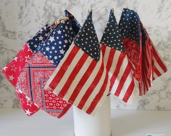 May include: A collection of small fabric flags featuring red, white, and blue patterns, including stars, stripes, and paisley designs, attached to wooden sticks and arranged in a plain white cylindrical container. The text Nina's Artsy Mart is visible.