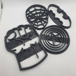 May include: Four black plastic cookie cutters shaped like superhero symbols: a fist, a spider web, a bat, and a star inside a circle.