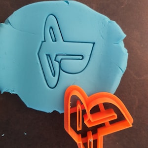 May include: A close-up of an orange plastic cookie cutter shaped like the PlayStation logo. The cookie cutter is sitting on top of a blue piece of modeling clay.
