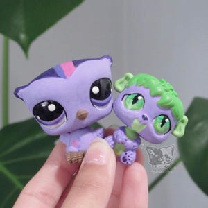 May include: Two small plastic animal figurines. The left figure is a purple owl with large black eyes, a brown beak, and a pink star detail. The right figure is a purple creature with green hair, green eyes, and a small paw print on its foot.