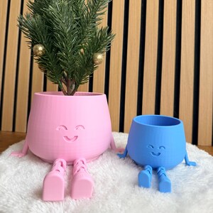 Könnte beinhalten: Zwei skurrile Pflanzgefäße, eines rosa und eines blauen, jeweils mit einem lächelnden Gesicht, Armen und Beinen. Der rosa Pflanztopf enthält einen kleinen immergrünen Baum, der mit goldenen Kugeln geschmückt ist. Die Pflanzgefäße stehen auf einer weißen, flauschigen Oberfläche.