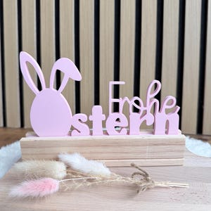 Könnte beinhalten: Rosa Osterdekoration aus Holz mit Hasenohren-Silhouette und dem deutschen Schriftzug "Frohe Ostern". Die Dekoration steht auf einem Holzsockel mit Trockenblumen vor einem Hintergrund aus Holzpaneelen.