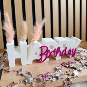 Könnte beinhalten: Ein weißer "HAPPY BIRTHDAY" Tortenaufsatz mit rosa Akzenten und Trockenblumen. Das Wort "HAPPY" ist in Blockbuchstaben und "Birthday" in Schreibschrift. Eine silberne Sternengirlande ist um die Basis drapiert. Der Hintergrund ist eine Holzoberfläche.