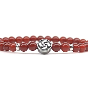 May include: Red beaded bracelet with a silver-colored Basque cross charm. The bracelet features small, round, polished red beads and silver-colored spacer beads. The charm is a stylized, four-armed design.