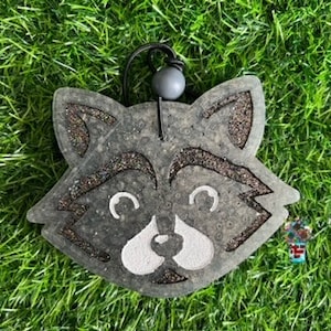 May include: A gray raccoon-shaped air freshener with a black string and a gray bead. The raccoon has white eyes, nose, and mouth.