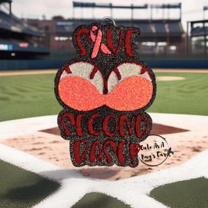 Peut inclure: Un ruban rose avec les mots "Save Second Base!" en paillettes rouges sur un fond noir. L'arrière-plan est un terrain de baseball.