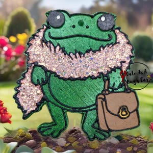 May include: A green frog wearing a pink sparkly scarf and carrying a brown purse. The frog is standing on a bed of green moss and dirt.
