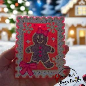 May include: A festive, rectangular ornament featuring a gingerbread man design. The ornament has a red background with a gingerbread man wearing a bow, surrounded by snowflakes. The border is white with glitter accents. The ornament is held in a hand.