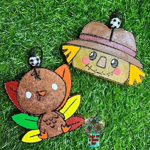 May include: Two felt ornaments, a turkey and a scarecrow, with black and white checkered accents. The turkey is brown with a red beak and yellow and green feathers. The scarecrow is brown with a straw hat and a yellow and brown face.