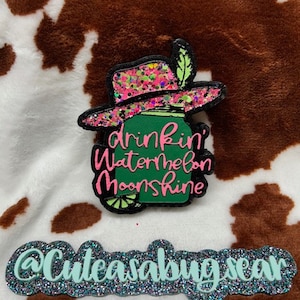 May include: A green mason jar with a pink glitter cowboy hat and the text "drinkin' watermelon moonshine". The jar is decorated with a lime slice and a green leaf. The image is on a brown and white cow print background. The text "@Cuteasabugsear" is at the bottom of the image.