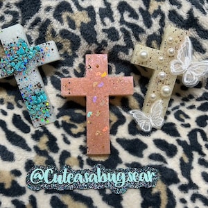 May include: Three resin crosses with different embellishments. One cross is white with turquoise glitter and stones, another is pink with white and gold glitter, and the last is white with pearl beads and a white butterfly.