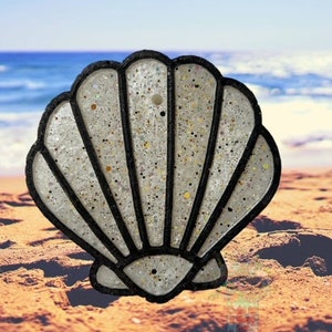 May include: A white seashell with black outlines, made of resin and glitter, is sitting on a sandy beach. The seashell has a detailed design with multiple ridges.