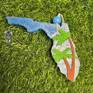 May include: A blue and white outline of the state of Florida with a brown palm tree and green leaves. The ornament has a blue bead and white string for hanging.