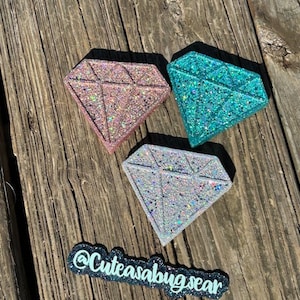 May include: Three glitter diamond-shaped coasters in pink, teal, and silver. The coasters are on a wooden surface. The text "@Cuteasabugsear" is visible in the bottom right corner of the image.