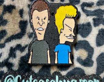 May include: A black and white cartoon character figurine of two men with brown and blonde hair. The figurine is on a leopard print background. The text "@Cuteasabugsear" is in the foreground.