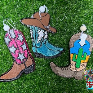May include: Three Christmas ornaments shaped like cowboy boots. One boot is blue with a white star design, one is brown with pink and white stars, and one is brown with a green cactus design.