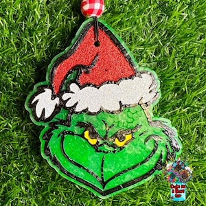 May include: Green Grinch ornament with a red and white Santa hat. The Grinch is wearing a white fur trim around his hat. The ornament is made of resin and has a glittery finish.