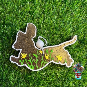 Könnte beinhalten: Ein brauner und goldener Dackel-Anhänger mit gelben Blumen und grünen Blättern. Der Anhänger hat eine weiße Schnur mit einer weißen Perle.