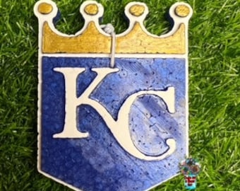 Può includere: Un ornamento in ceramica blu e bianca con una corona dorata e le lettere "KC" in bianco. L'ornamento ha la forma di uno scudo.