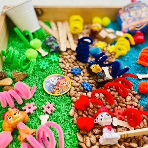 May include: A wooden tray filled with sensory play items. The tray contains green dyed rice, beans, and blue dyed rice. There are toy figures, pipe cleaners, wooden sticks, and other colorful objects. A Paw Patrol logo is visible.