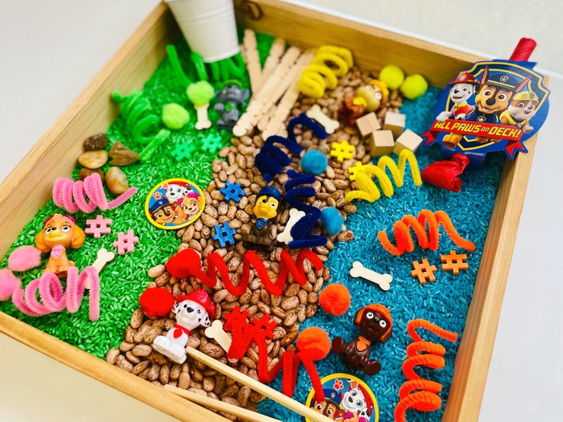 May include: A wooden tray filled with sensory materials, including green rice, pinto beans, and blue rice. The tray features Paw Patrol character figurines, pipe cleaners, wooden blocks, and a badge that reads "ALL PAWS ON DECK!"