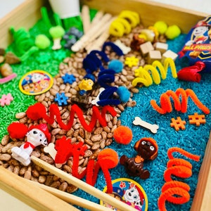 May include: A wooden tray filled with a sensory bin featuring a Paw Patrol theme. The bin includes green and blue dyed rice, beans, toy figures, pipe cleaners, wooden sticks, and a card that reads "ALL PAWS ON DECK!"