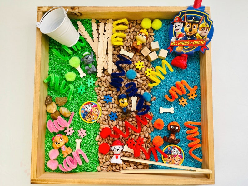 May include: A wooden tray filled with various sensory items. Includes green and blue dyed rice, beans, small toys, pipe cleaners, wooden blocks, and Paw Patrol character figures. The tray also contains a small white bucket and a Paw Patrol badge that reads "All Paws on Deck!"