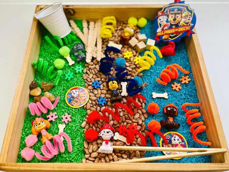 May include: A wooden tray filled with sensory play items. Includes green and blue dyed rice, beans, pipe cleaners, small toys, wooden blocks, and a Paw Patrol badge. The tray is designed for children's play and exploration.