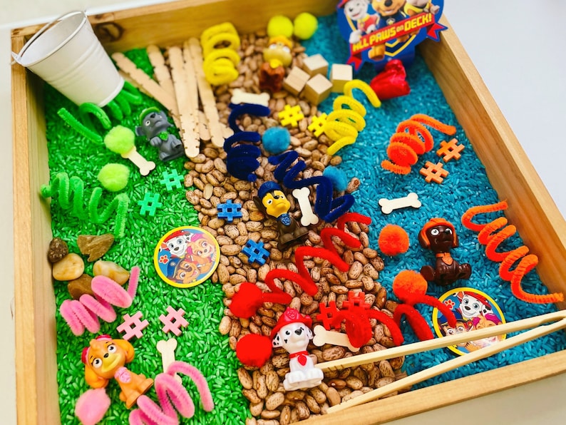 May include: A wooden tray filled with sensory play items. Includes green and blue dyed rice, beans, toy figures, pipe cleaners, wooden blocks, and small accessories. Features a Paw Patrol theme with character figures and logo.