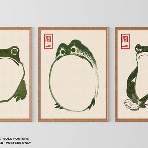 Op de afbeelding: Drie ingelijste prints van groene kikkers in verschillende poses. De eerste kikker zit met gekruiste benen. De tweede kikker is rond. De derde kikker houdt een kom en een stok vast. Elke print heeft een rode stempel met het Japanse karakter "開" in de linkerbovenhoek.