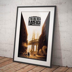 Può includere: Poster incorniciato con la scritta "C'era Una Volta In America". L'immagine mostra un ponte e delle figure su una strada, incorniciata da un bordo nero. Il poster è appoggiato a un muro strutturato. Un articolo a tema cinematografico.