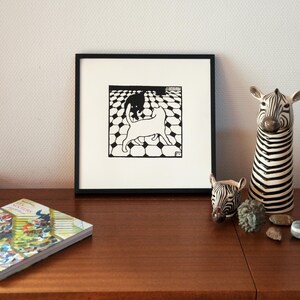 K&ouml;nnte beinhalten: Ein schwarz-wei&szlig;er gerahmter Druck von zwei Katzen, die auf einem karierten Boden spielen. Der Druck h&auml;ngt an einer Wand &uuml;ber einem Holzregal mit B&uuml;chern und dekorativen Zebrafiguren.