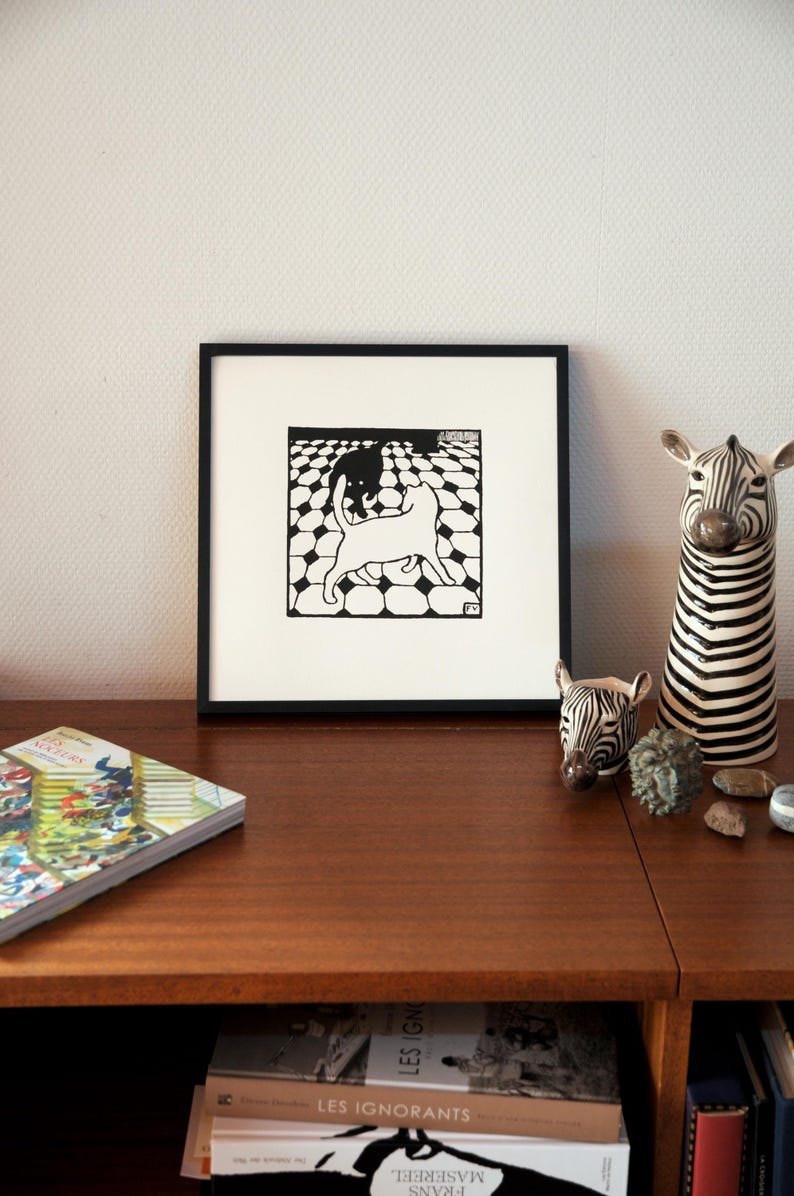 K&ouml;nnte beinhalten: Ein schwarz-wei&szlig;er gerahmter Druck von zwei Katzen, die auf einem karierten Boden spielen. Der Druck h&auml;ngt an einer Wand &uuml;ber einem Holzregal mit B&uuml;chern und dekorativen Zebrafiguren.