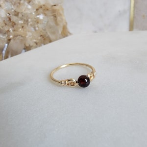 May include: A gold-colored ring with a dark red gemstone. The ring features a delicate wire design, with the gemstone centered and secured by wrapped wire. The ring is set against a white background.