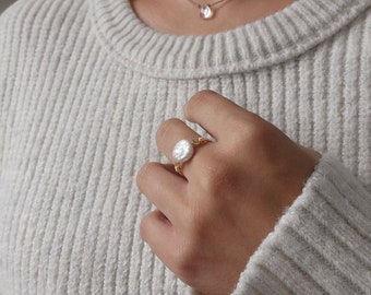 May include: A gold ring with a large, round, white pearl. The ring is on a person's finger.