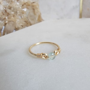 May include: A delicate gold ring with a light blue gemstone. The ring is made with a simple wire design and features a single round gemstone.