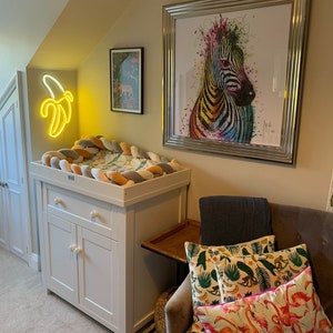 May include: A white changing table with a drawer and two doors. The changing pad is covered with a multi-colored braided fabric. A neon yellow banana shaped light is on the wall behind the changing table. A framed print of a colorful zebra is on the wall above the changing table. A chair with a gray throw blanket and colorful pillows is in the corner of the room.