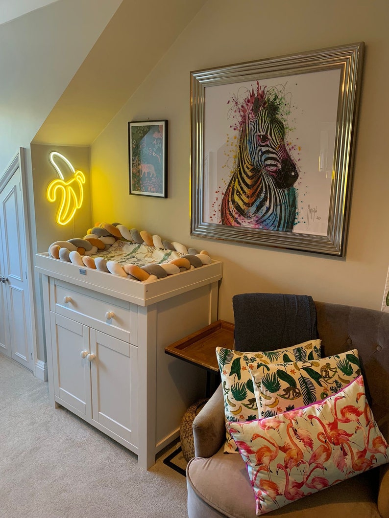 May include: A white changing table with a drawer and two doors. The changing pad is covered with a multi-colored braided fabric. A neon yellow banana shaped light is on the wall behind the changing table. A framed print of a colorful zebra is on the wall above the changing table. A chair with a gray throw blanket and colorful pillows is in the corner of the room.