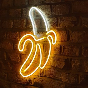 May include: A neon sign shaped like a banana, with a yellow glow against a brick wall background.