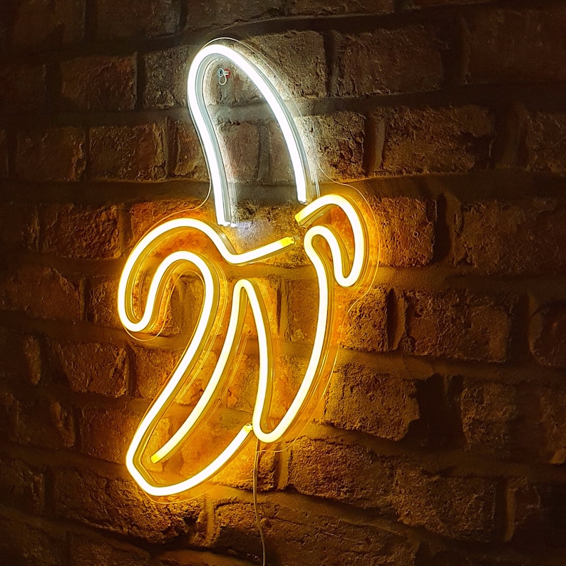 May include: A neon sign shaped like a banana, with a yellow glow against a brick wall background.
