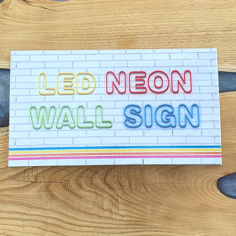 May include: A white brick wall sign with the text "LED NEON WALL SIGN" in colorful neon letters. The letters are outlined in black and have a 3D effect. The sign has a rainbow stripe border at the bottom.