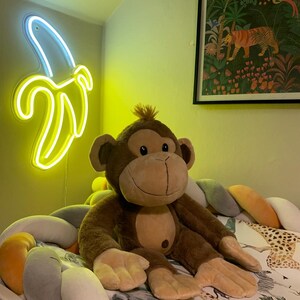 May include: A brown plush monkey toy sitting on a white and gray braided bed bumper. The bumper is decorated with a yellow, orange, and gray pattern. A neon yellow banana-shaped light is on the wall behind the monkey.