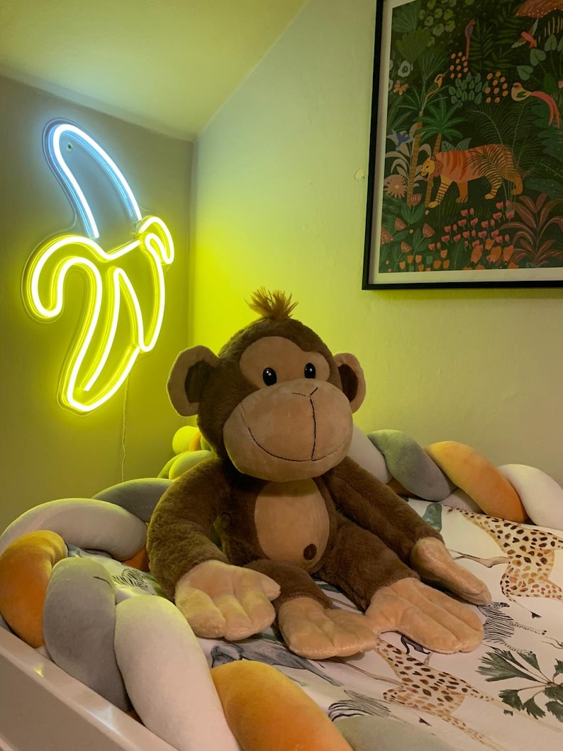 May include: A brown plush monkey toy sitting on a white and gray braided bed bumper. The bumper is decorated with a yellow, orange, and gray pattern. A neon yellow banana-shaped light is on the wall behind the monkey.
