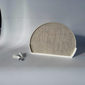 May include: A semi-circular, neutral-toned wall decor piece with a textured fabric insert, set in a white frame. Includes two white wall anchors and a screw, suggesting easy installation. The design is minimalist and modern.